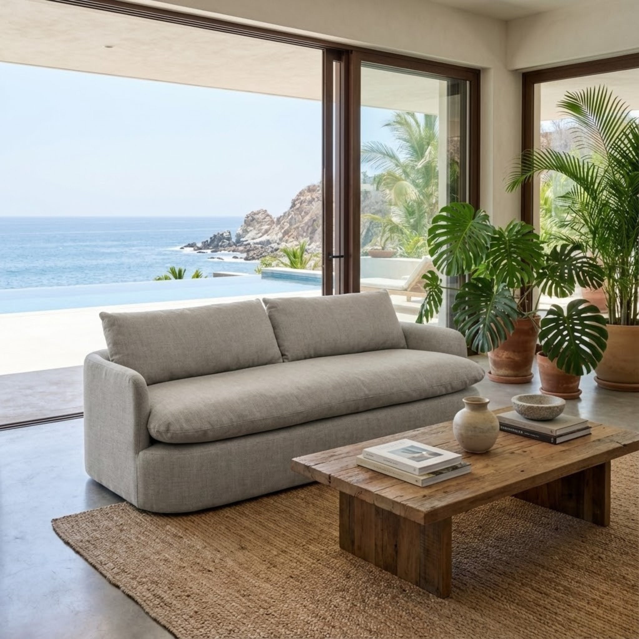 Sofá Sereno Stone en una sala de lujo frente al mar, con decoración tropical y mesa de madera rústica.